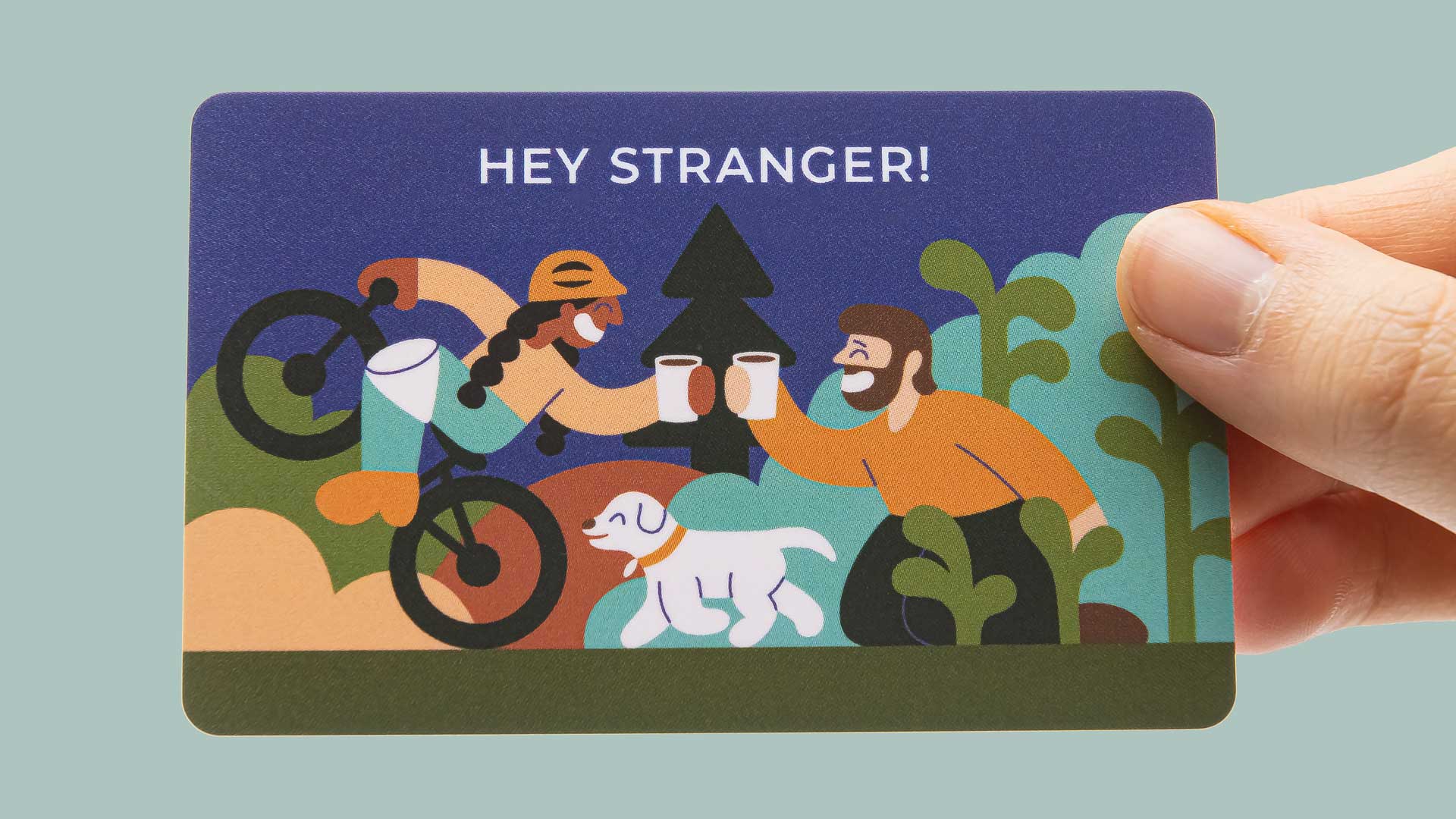 An image of a gift card held by a person’s hand. The gift card has an illustration of a brown woman on a mountain bike, a white dog, and a white man with a beard. The two people clink coffees.
