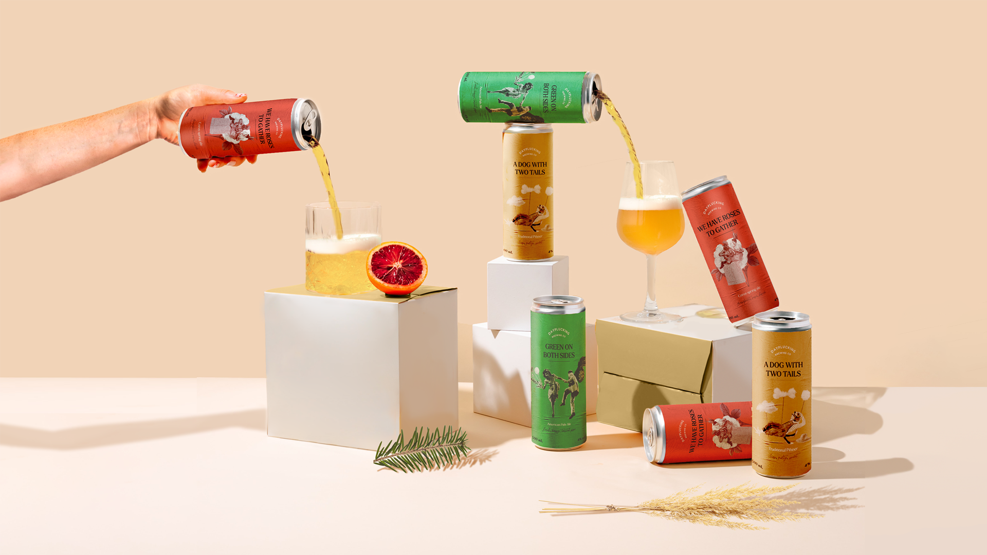 An image of 3 different beers on a set of white boxes staggering in height. The background of the set is beige, while the cans alternate between red, green, and brown, each with different assigned flavors. A hand pours beer into a glass on the left side of the image. A blood orange, a pine sprig, and a wheat bundle are scattered around the set next to their matching cans.