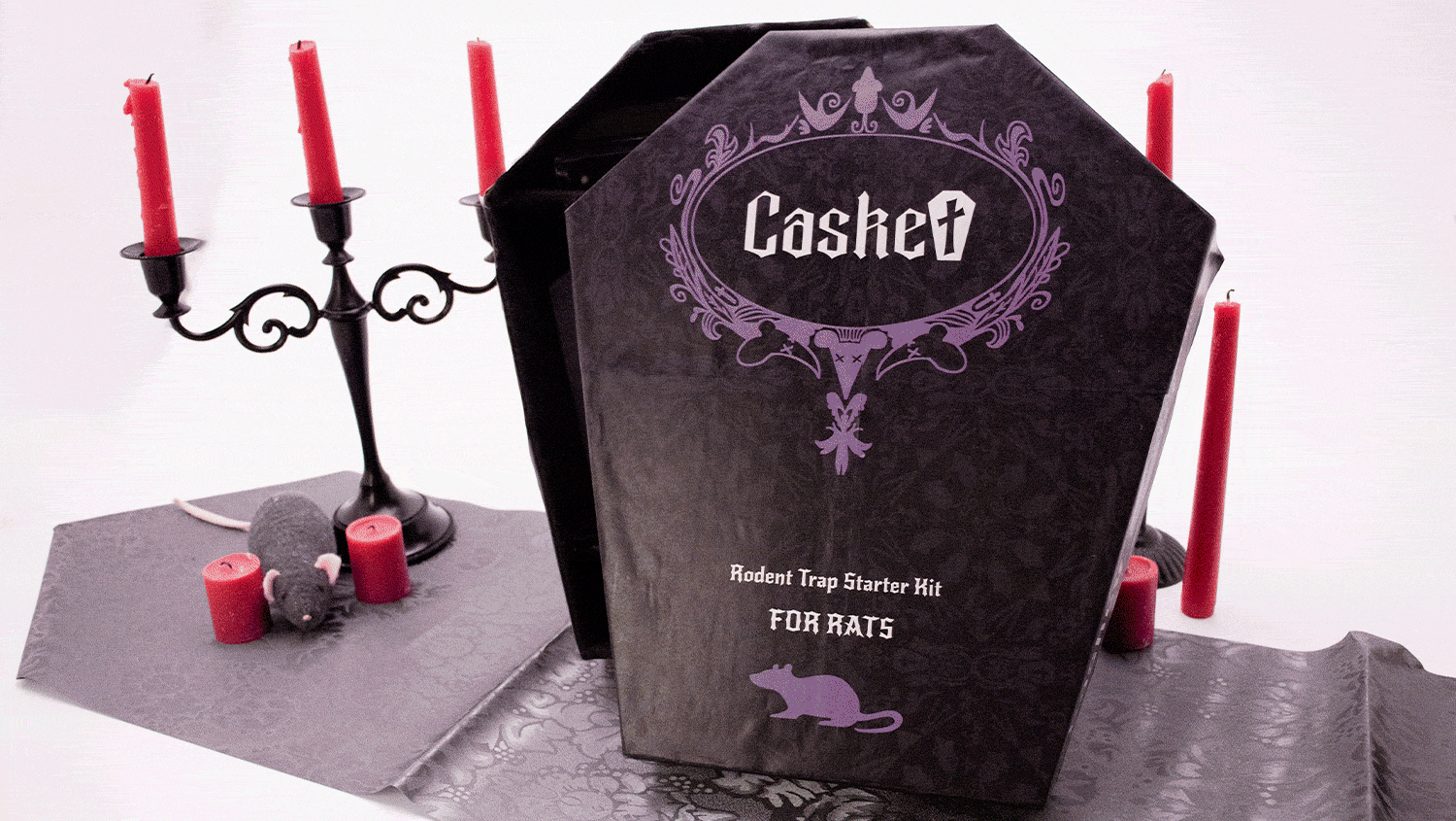 Looping GIF: a black coffin-shaped box with Gothic text “Casket: Rodent Trap Starter Kit FOR RATS” amid red candles in black iron fixtures with a plush rat; in the same setting the inside of the box cover with the eulogy “In Loving Remembrance of Rat” is displayed; then, a black coffin-shaped box with compartments containing gothic-themed rat trapping paraphernalia; then, Gothic-themed rat trapping paraphernalia, including a coffin-shaped rat snap trap, disinfectant, bait, steel wool, disposable bags, and disposable gloves.